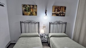 A twin room with two beds, a nightstand, a lamp, and two paintings on the walls. - Pensión Javier (Seville)