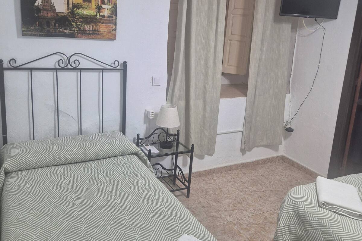 Habitación de hotel con cama, mesita de noche con lámpara, televisor montado en la pared y un cuadro de un edificio.