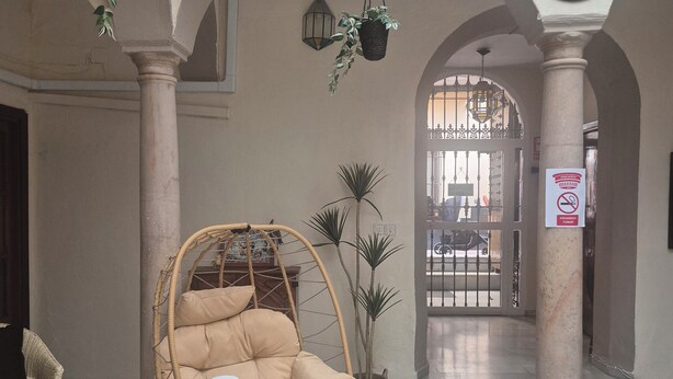 A spacious lobby with a hanging plant, a wicker chair, a tiled coffee table, and a decorative clock. - Pensión Javier (Seville)