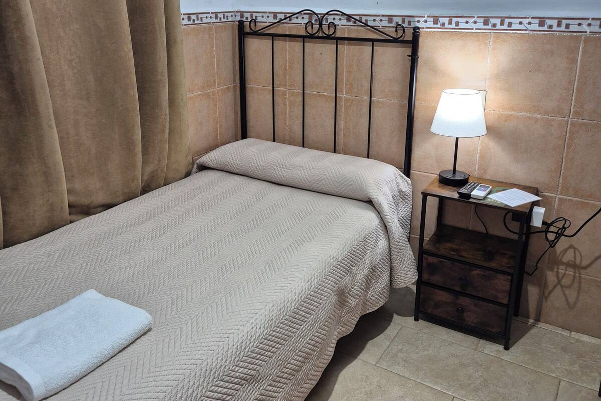Una cama individual con colcha color crema, una mesita de noche con lámpara y una cortina.