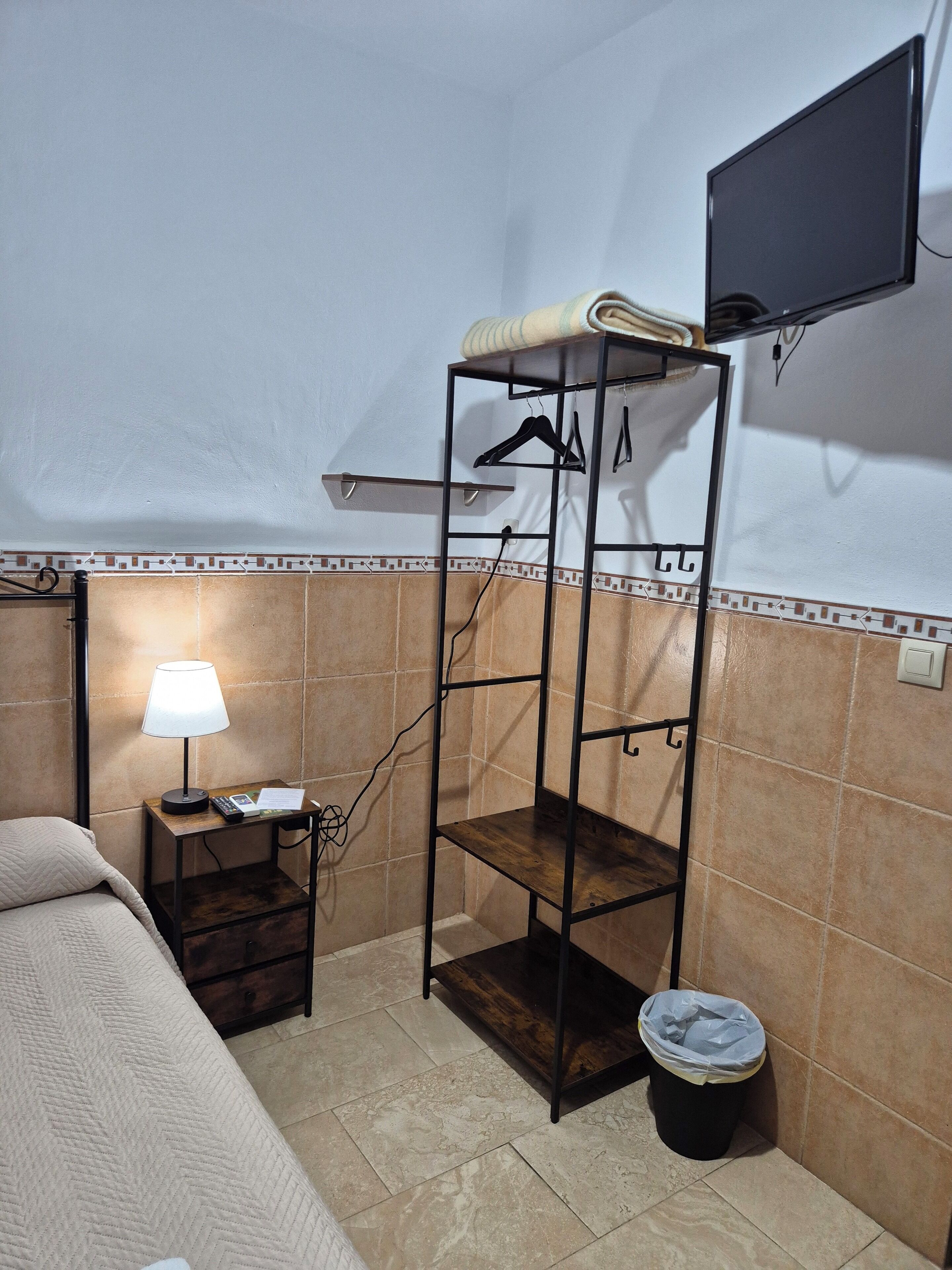 Una habitación pequeña y funcional con cama, mesita de noche con lámpara, televisor en la pared, perchero y cubo de basura.