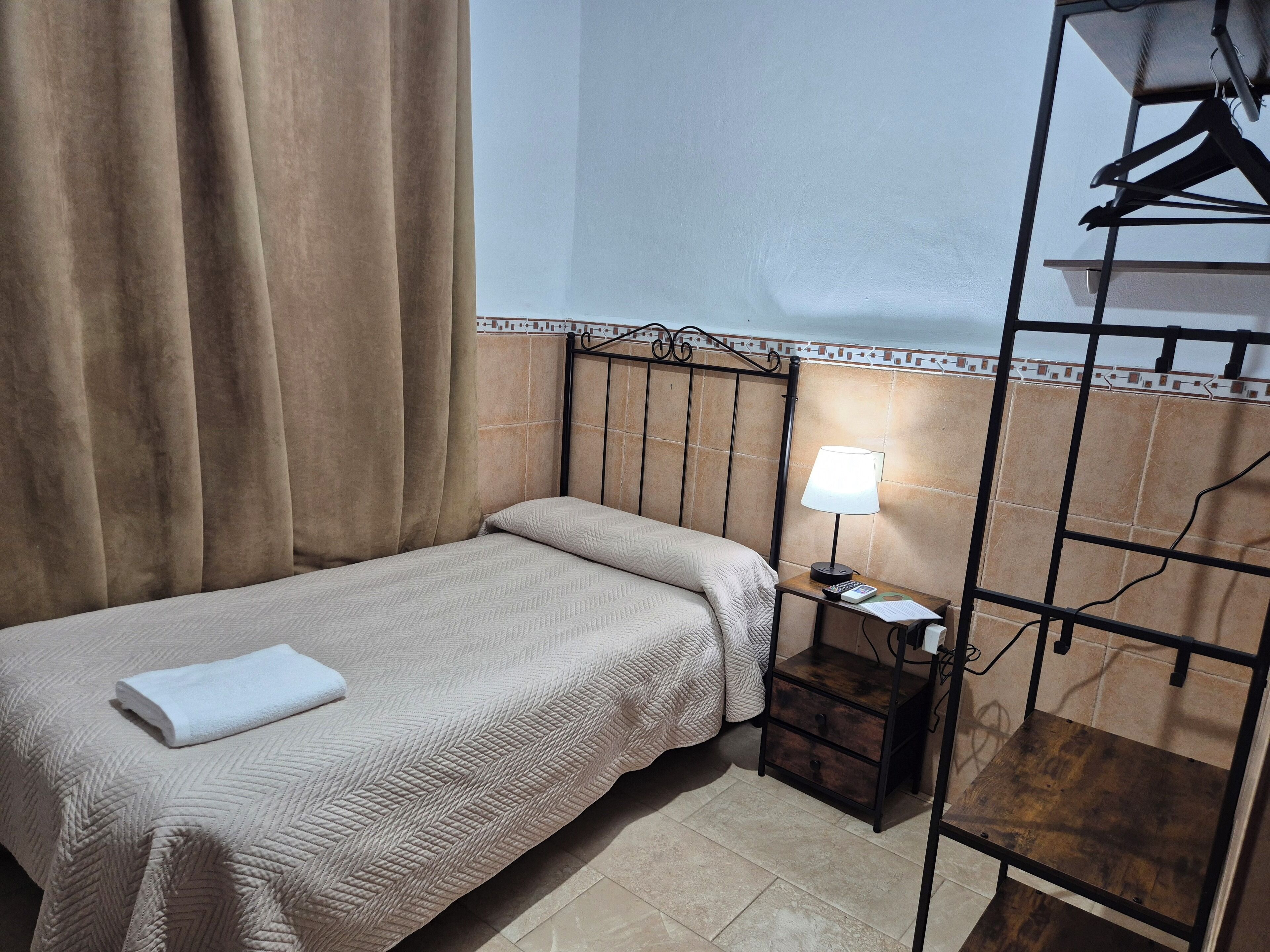 A single bed with a white towel, a nightstand with a lamp, and a wooden shelf.