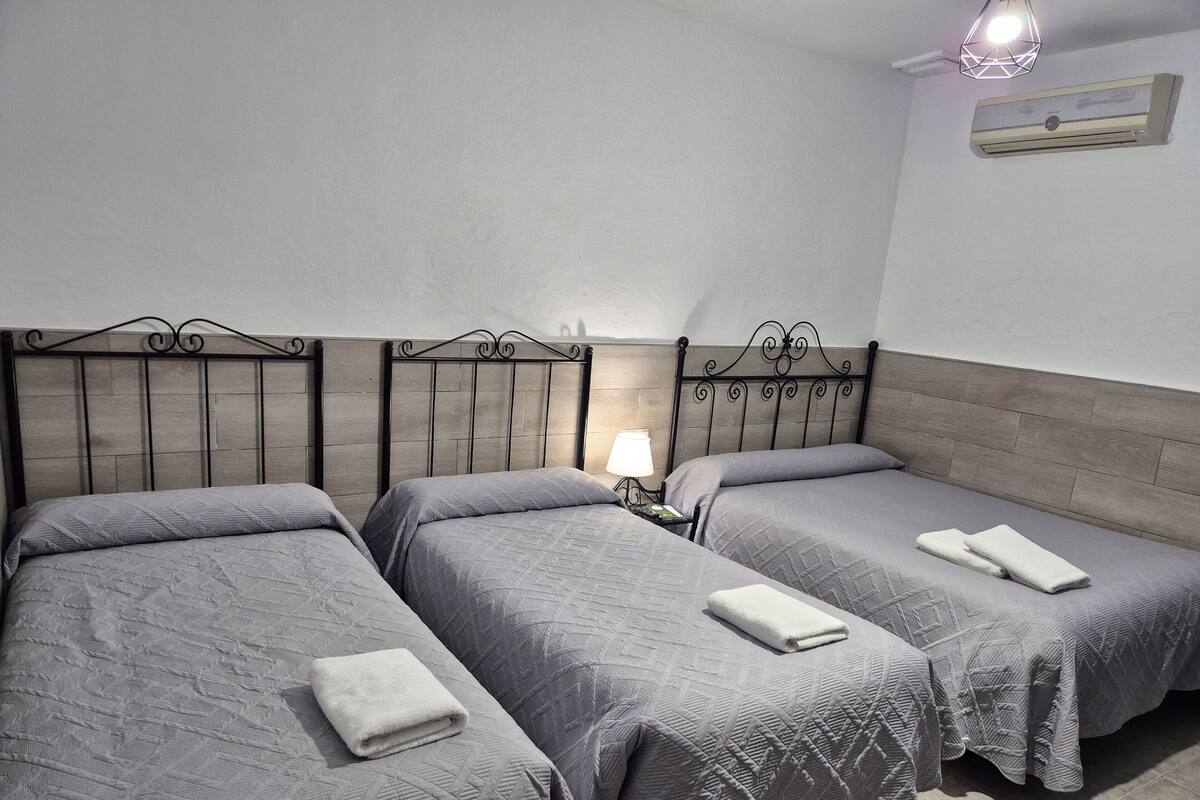 Habitación de hotel con tres camas, cada una con una colcha gris acolchada y una toalla blanca doblada. Se observa un aire acondicionado instalado en la pared y una lámpara de noche individual en la mesita.