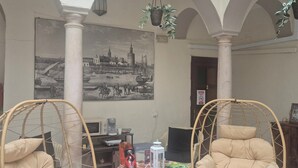 A spacious indoor area with high ceilings and arches, featuring two wicker chairs, a small round table with a coffee machine, and a large black-and-white cityscape painting on the wall. - Pensión Javier (Seville)