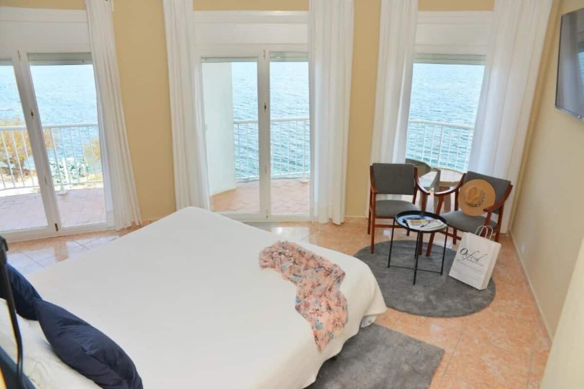 A hotel room with a bed, a small table with chairs, a view of the sea through large windows, and a balcony.