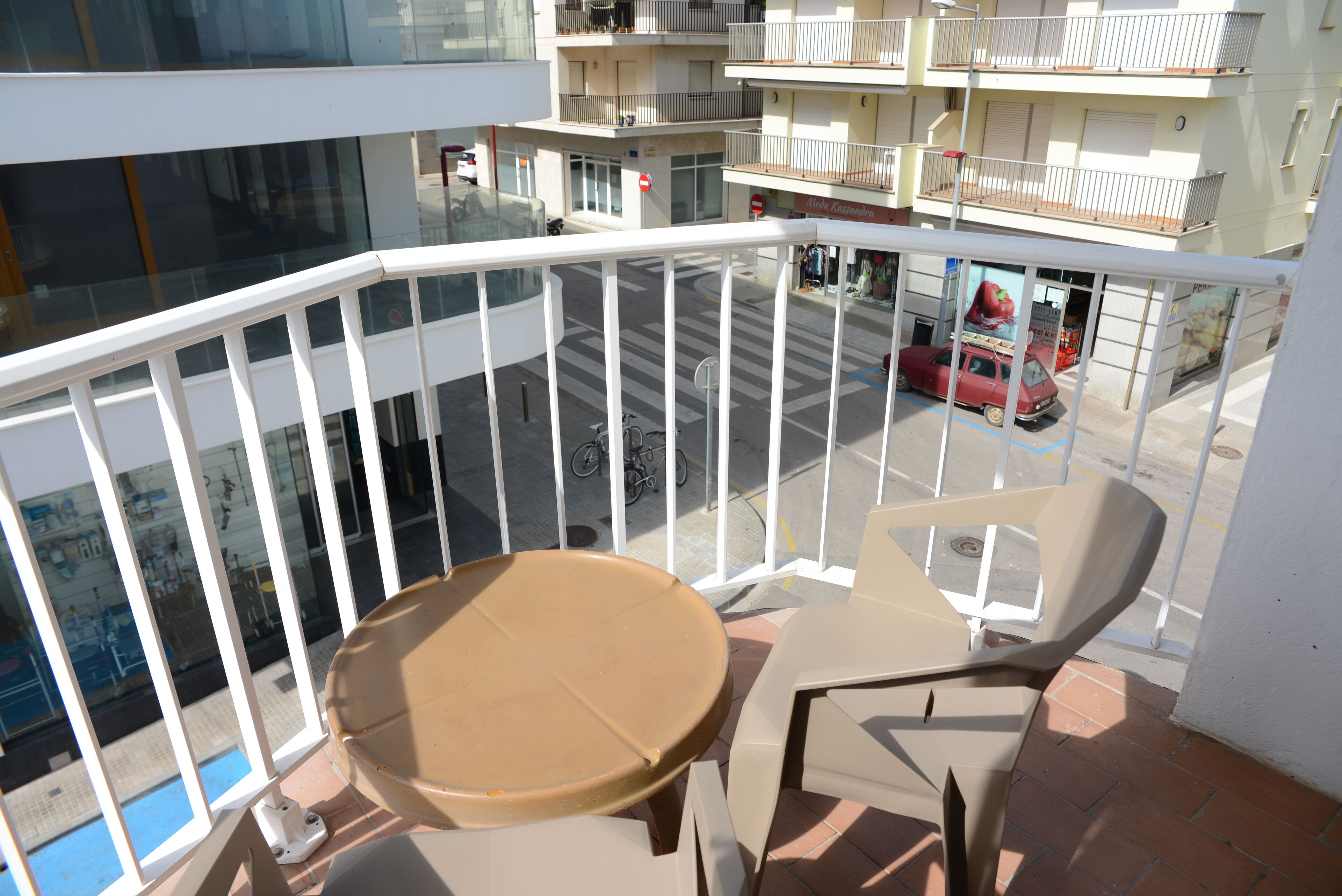 En balkon med et bord og stole, ud til en gade med en rød bil og en bygning med egne balkoner.