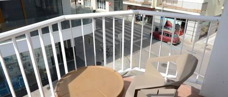 Een balkon met een tafel en stoelen, uitkijkend op een straat met een rode auto en een gebouw met balkons.