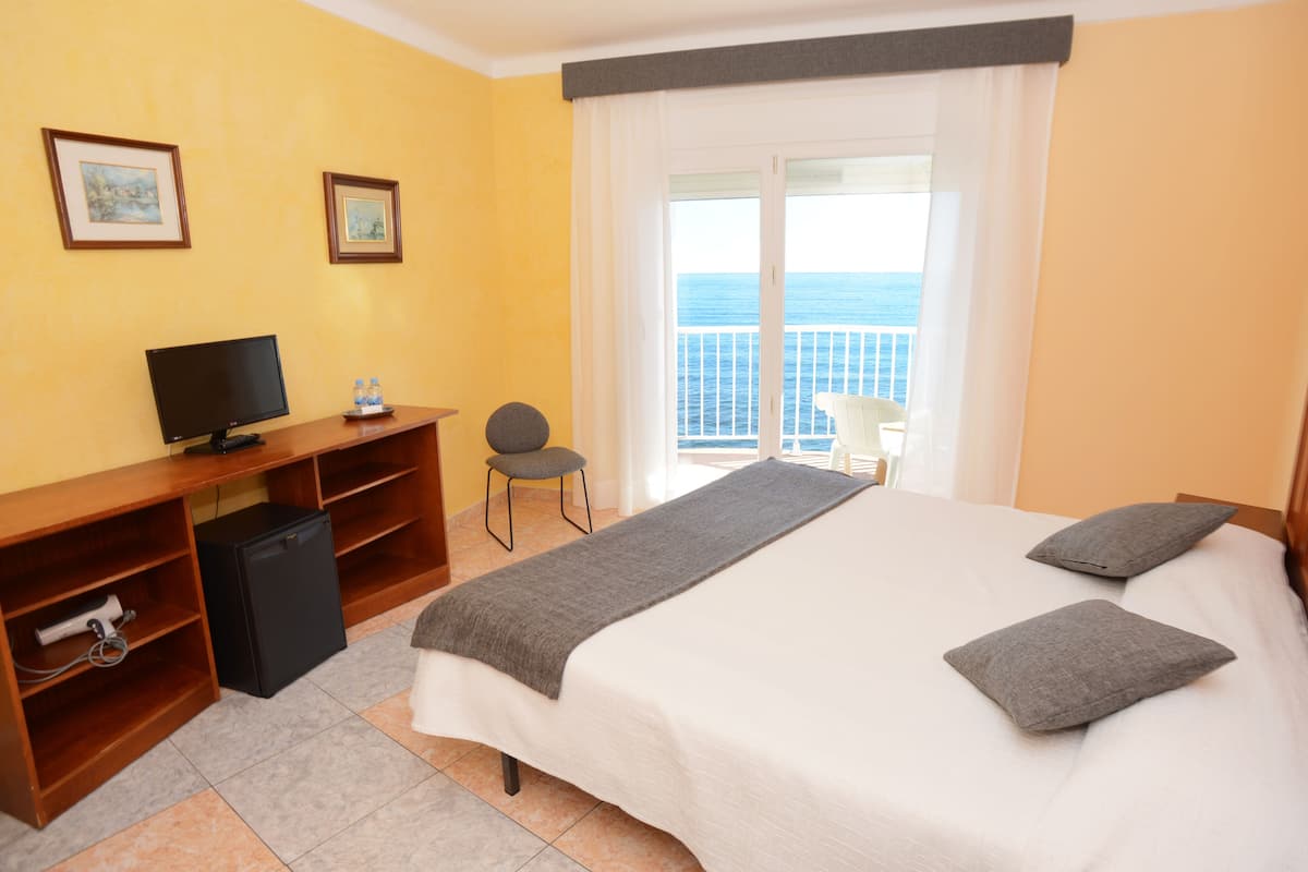 A hotel room with a bed, a desk with a computer, a chair, and a view of the sea through a balcony.