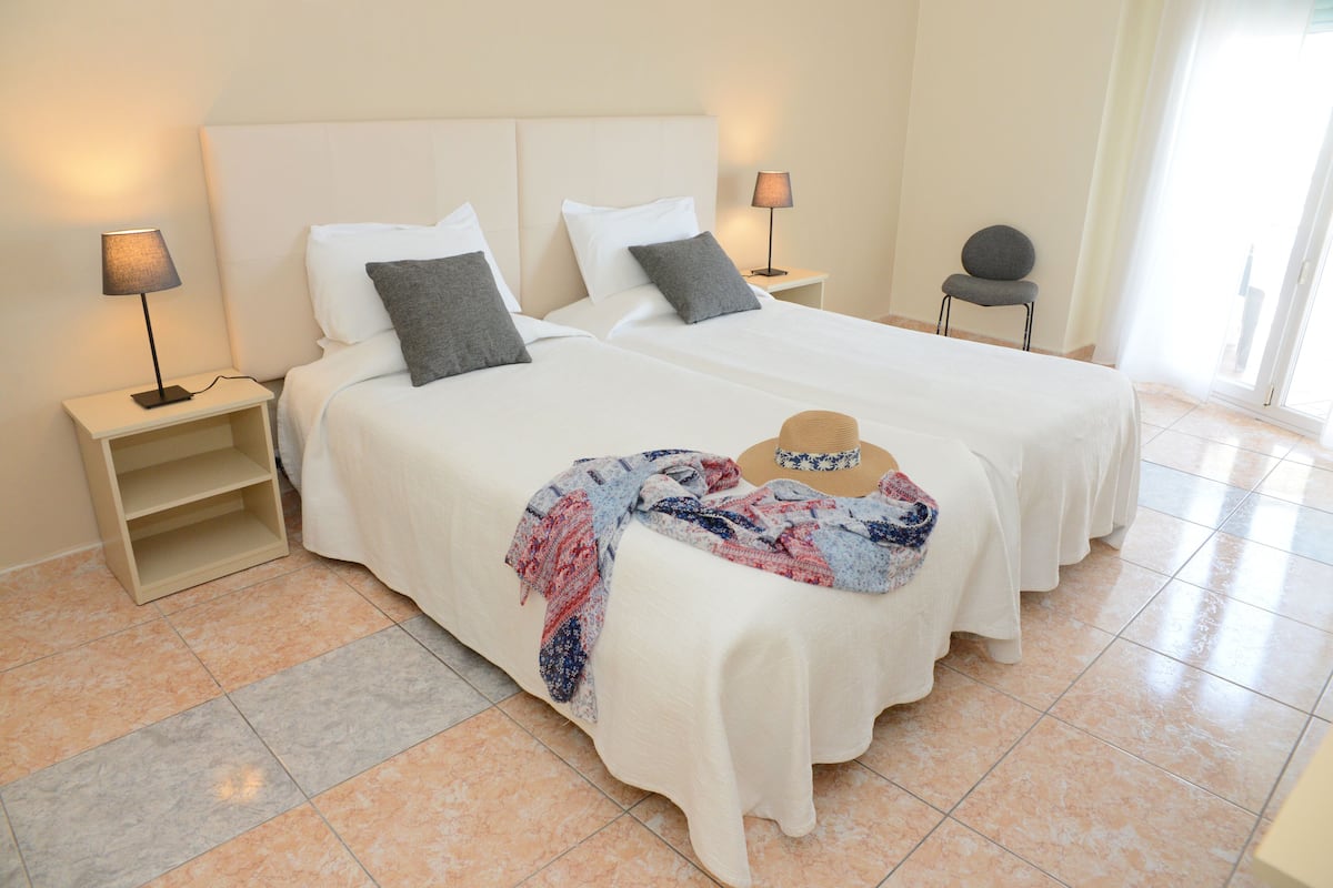 A hotel room with two beds, each with a white headboard, a bedside table with a lamp, and a chair.