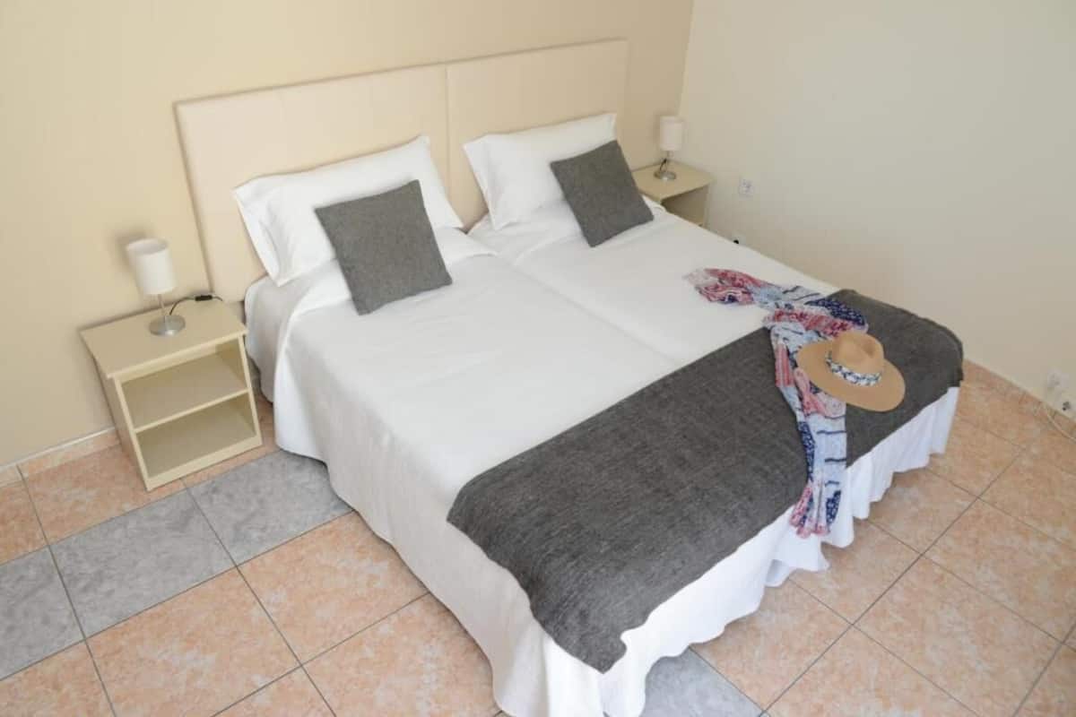 A double bed with white and gray bedding, a bedside table with a lamp, and a hat on the bed.