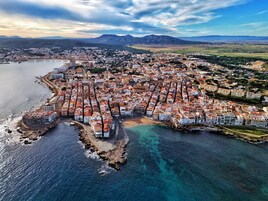 An aerial view of a coastal town with a dense cluster of buildings along the shoreline, a marina, and a mountainous backdrop.