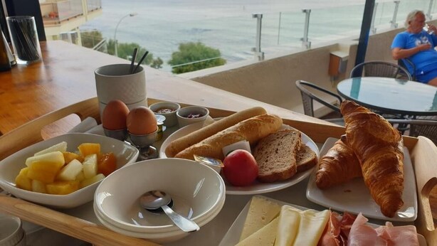 Bandeja de desayuno con cruasanes, queso, jamón y fruta en un balcón con vistas al mar.
