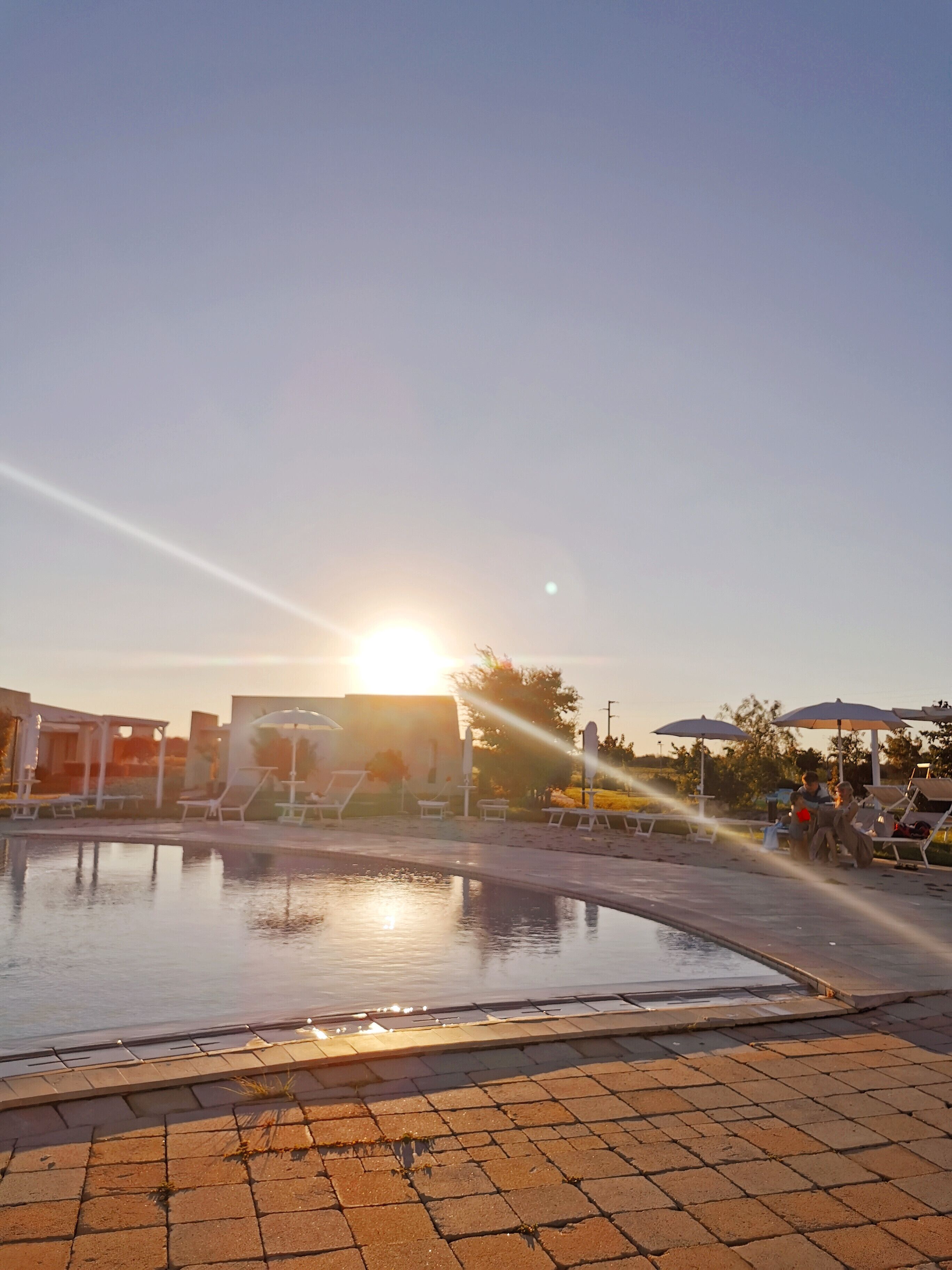 A swimming pool with lounge chairs and umbrellas, surrounded by buildings and trees.