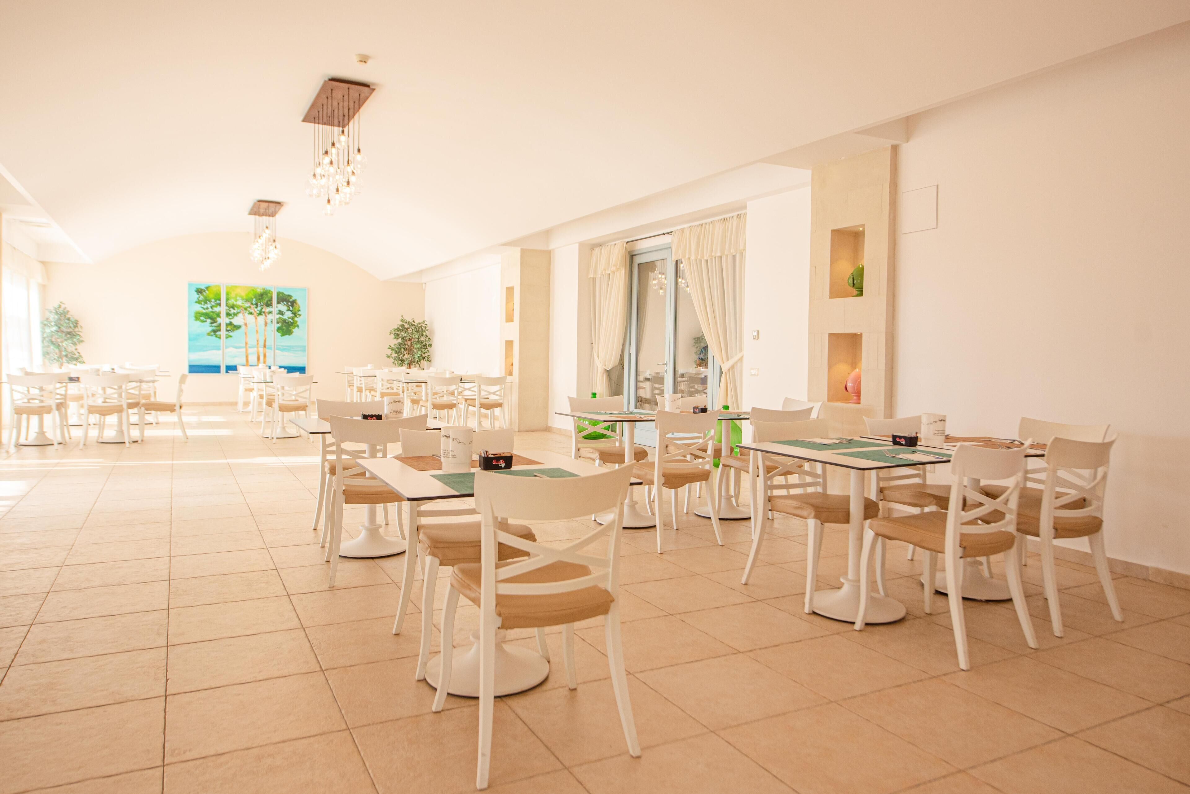 A spacious dining area with multiple tables and chairs, large windows with curtains, and a modern chandelier.