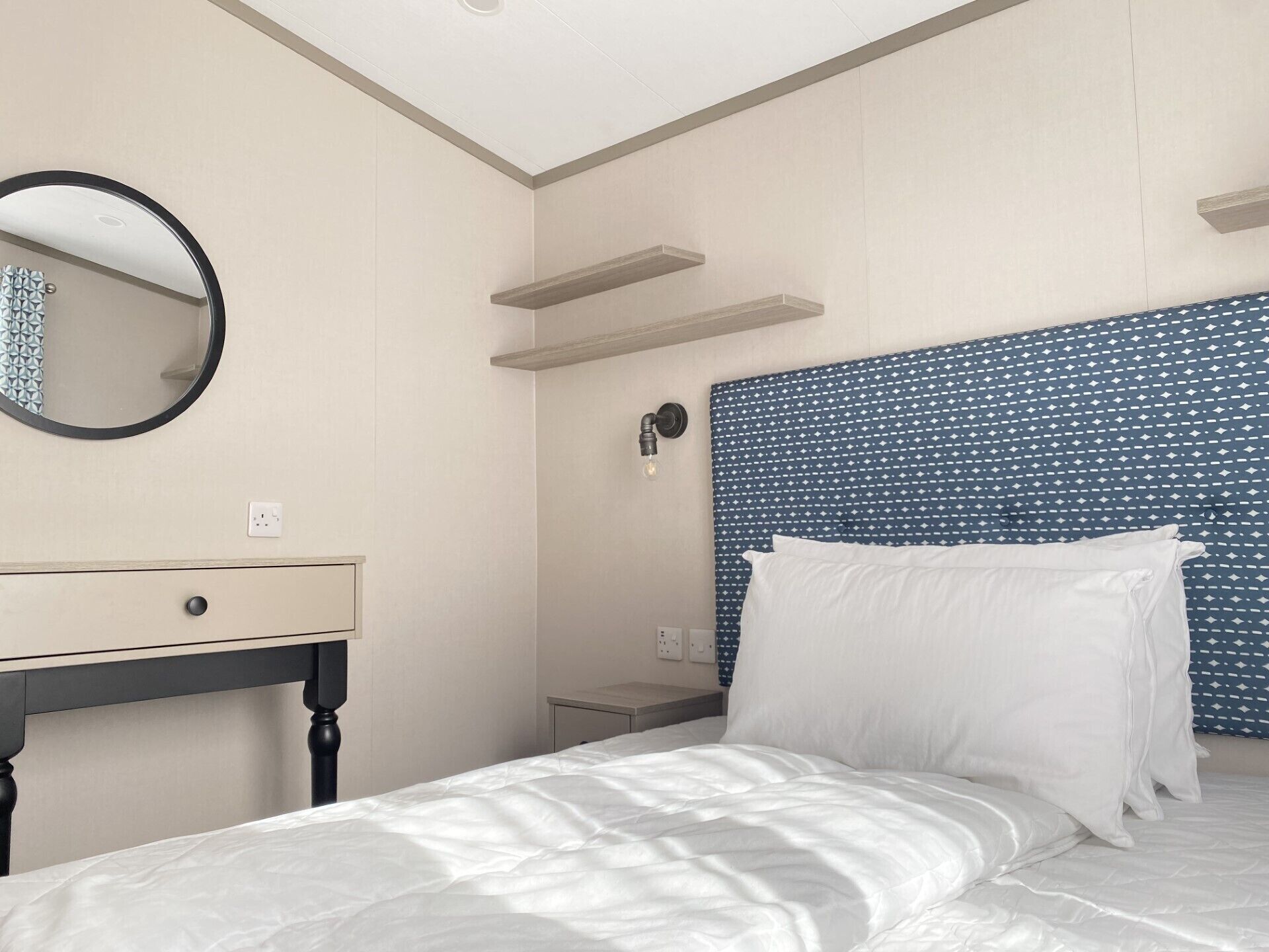 A bedroom with a bed, a bedside table, a mirror, and a wall-mounted shelf.