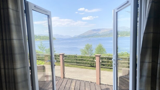 An open sliding glass door leading to a wooden deck with a view of a lake and mountains. - Linnhe Lochside Holidays (Fort William)