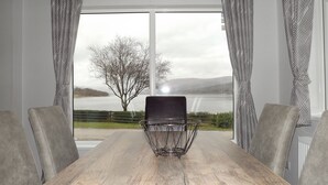 A dining area with a table and chairs, a metal chair with a black seat, and a view of a lake and tree through large windows. - Linnhe Lochside Holidays (Fort William)