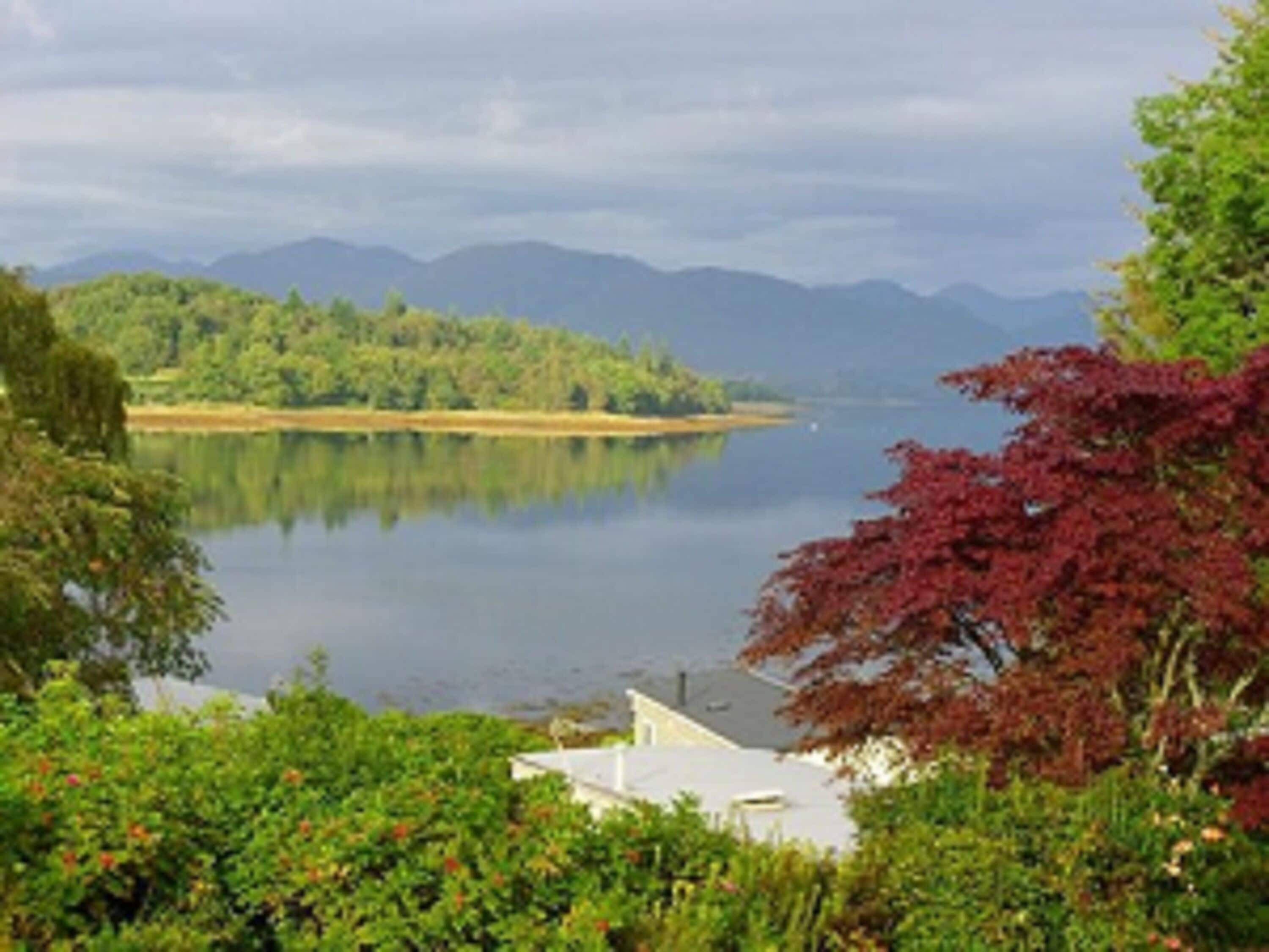 A serene lakeside view with a house in the foreground, surrounded by lush greenery and mountains in the background.