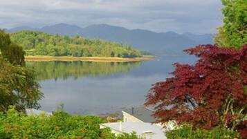 Une vue paisible sur un lac, avec une maison au premier plan, entourée d’une végétation luxuriante et des montagnes en arrière-plan.
