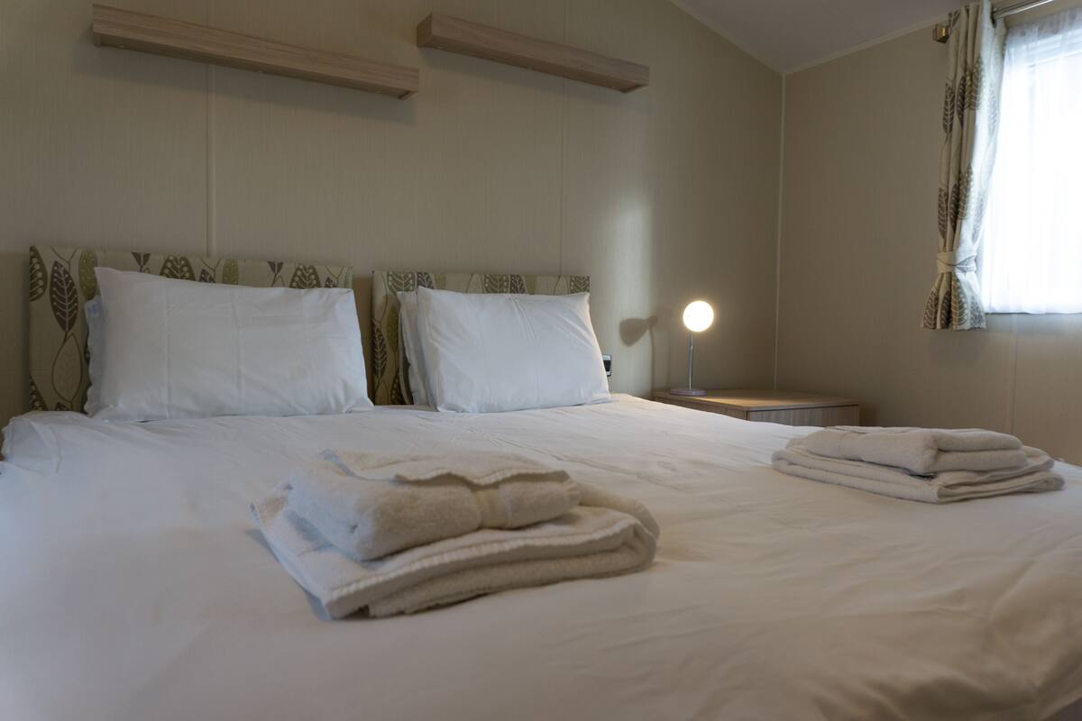 A neatly made bed with white linens and folded towels, a bedside lamp, and a window with curtains.