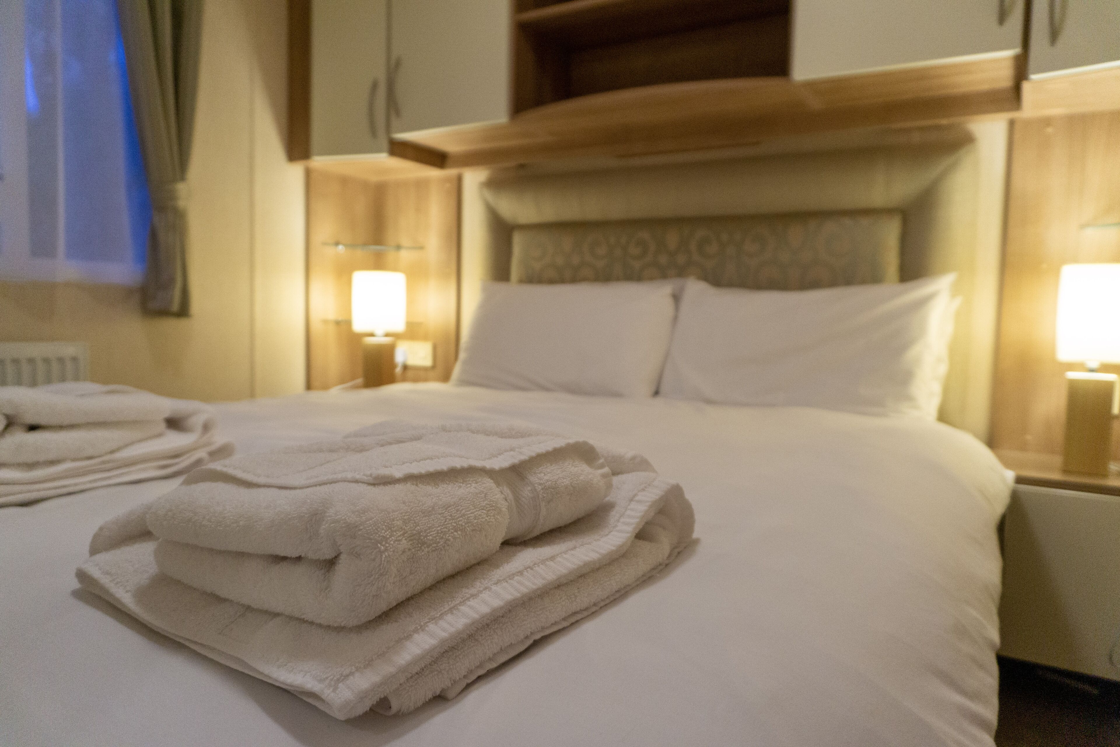 A neatly made bed with folded white towels, wooden headboard, and bedside lamps.