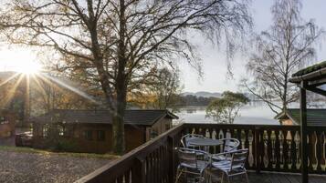Een houten terras met metalen stoelen met uitzicht op een meer, omgeven door bomen en een klein huisje.