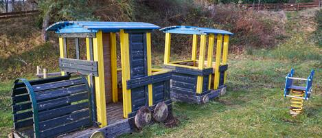 Een kleine, stilstaande trein met een blauw-geel kleurenschema, gelegen in een groene buitenruimte met bomen en een houten hek op de achtergrond.
