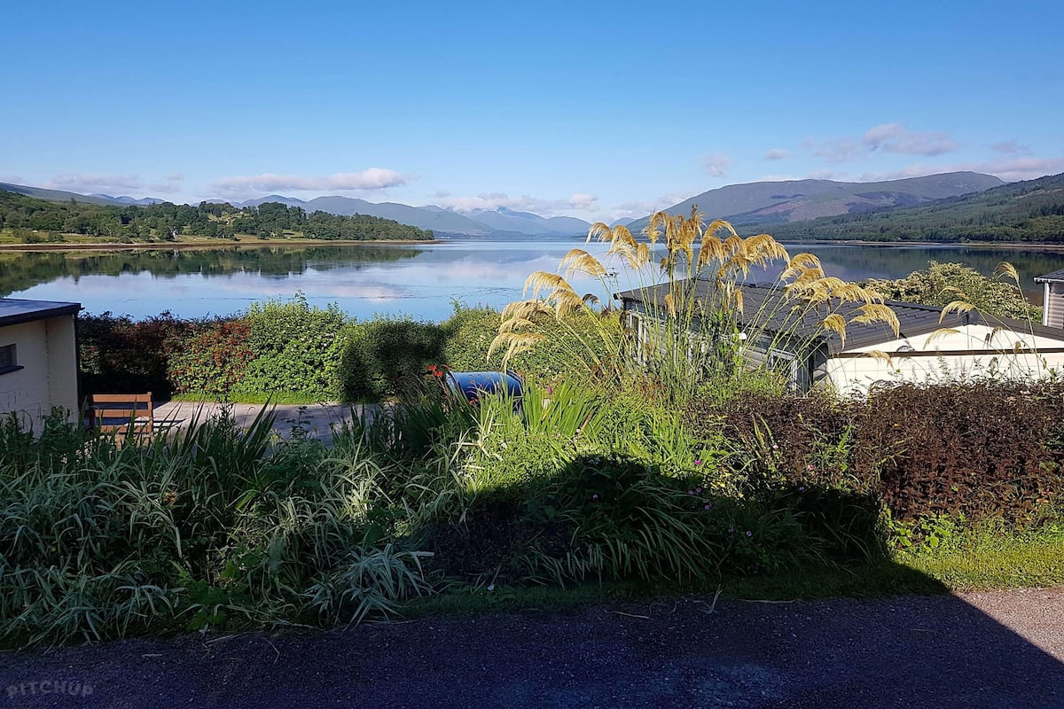A lakeside view with a clear sky, a calm lake, and surrounding greenery.