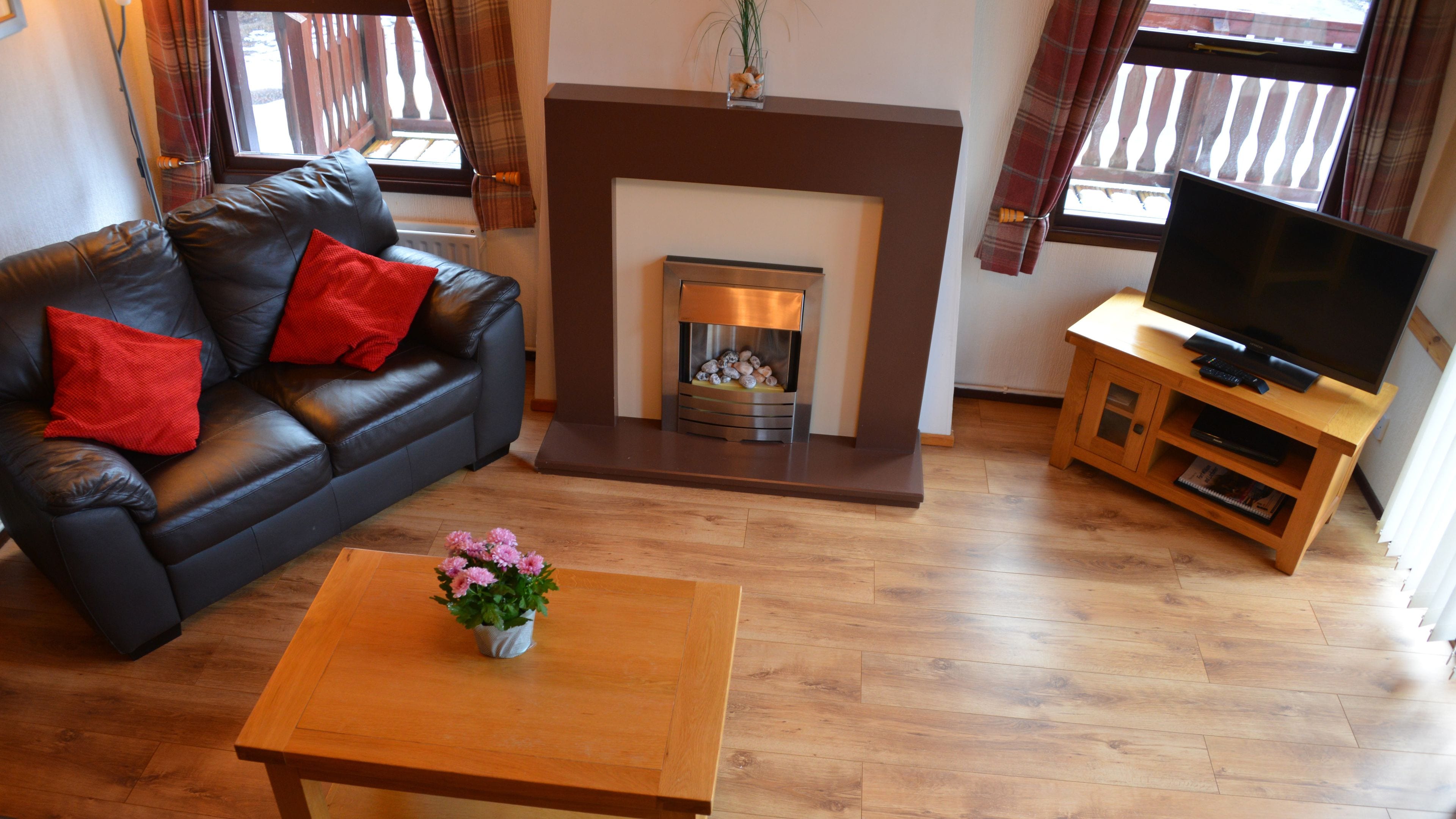 A living room with a fireplace, a sofa with red cushions, a wooden coffee table with a flower vase, and a TV on a wooden stand.