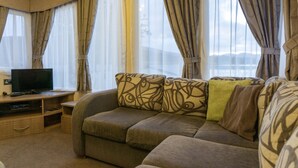 A living room with a sofa, a television on a wooden stand, and large windows with curtains. - Linnhe Lochside Holidays (Fort William)