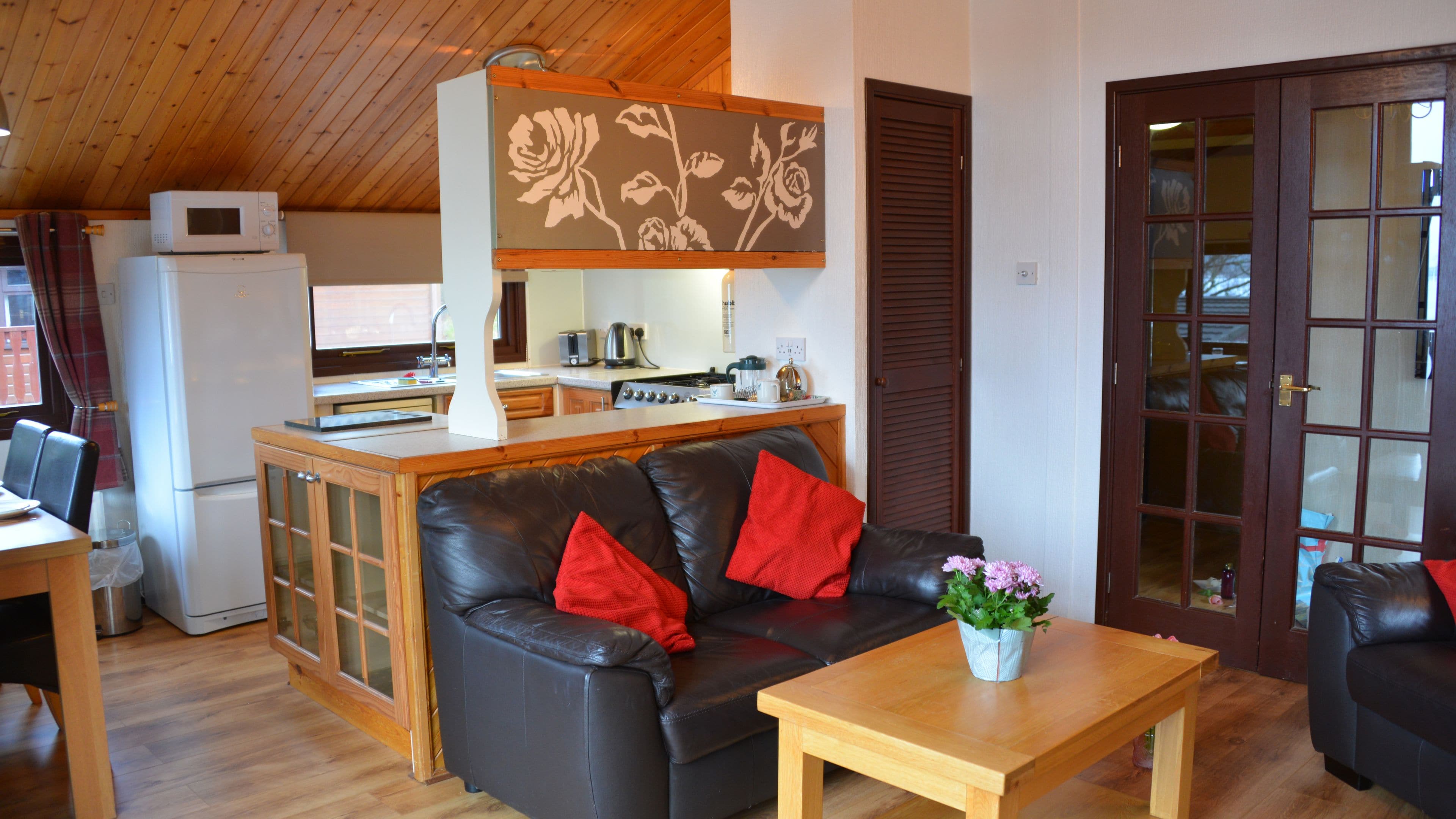 Un espacio habitable compacto con kitchenette, un sofá de cuero negro con cojines rojos, una mesa de centro de madera y una decoración floral en la pared.