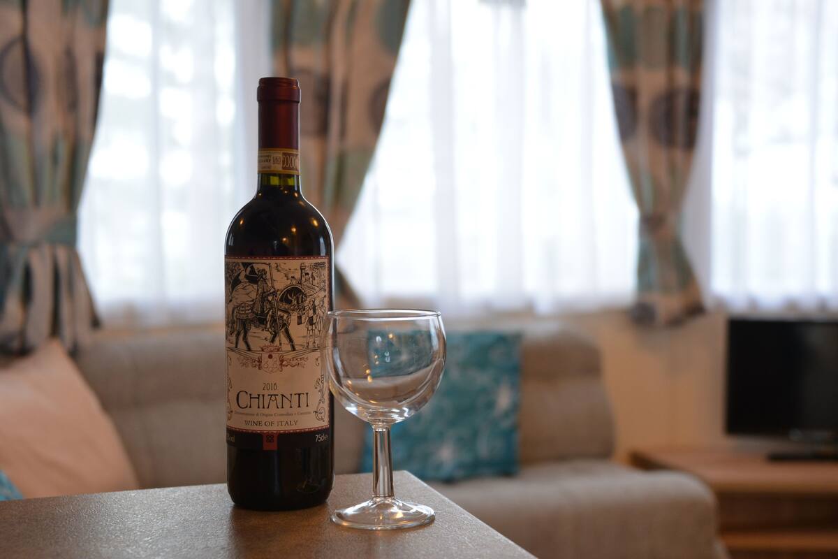 A bottle of Chianti wine and an empty glass on a table in a living room with a sofa and a TV in the background.