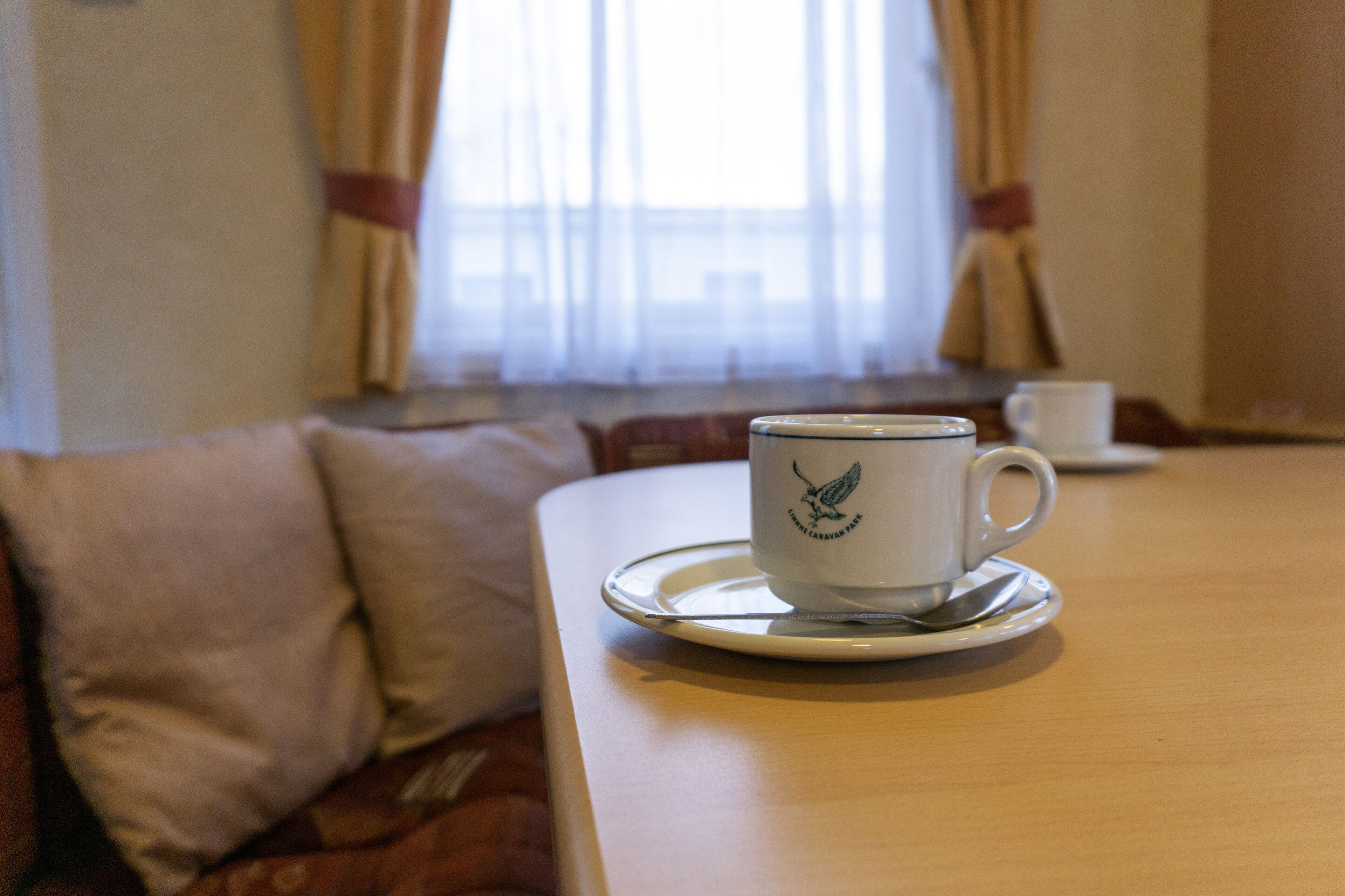 A cup of coffee on a saucer on a wooden table in a room with a sofa and a window with curtains.