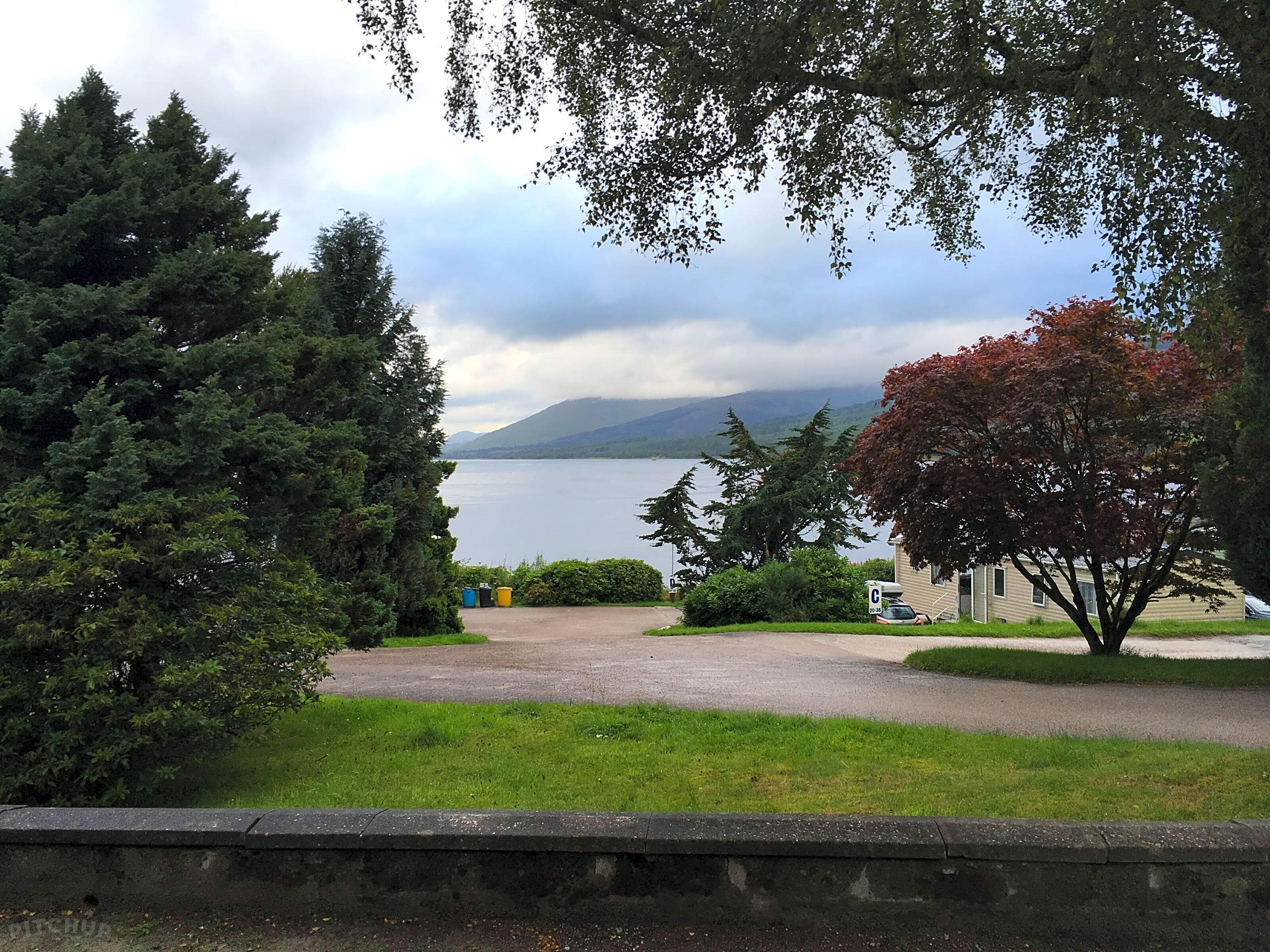 A view of a lake with mountains in the background, surrounded by trees and a parking area.