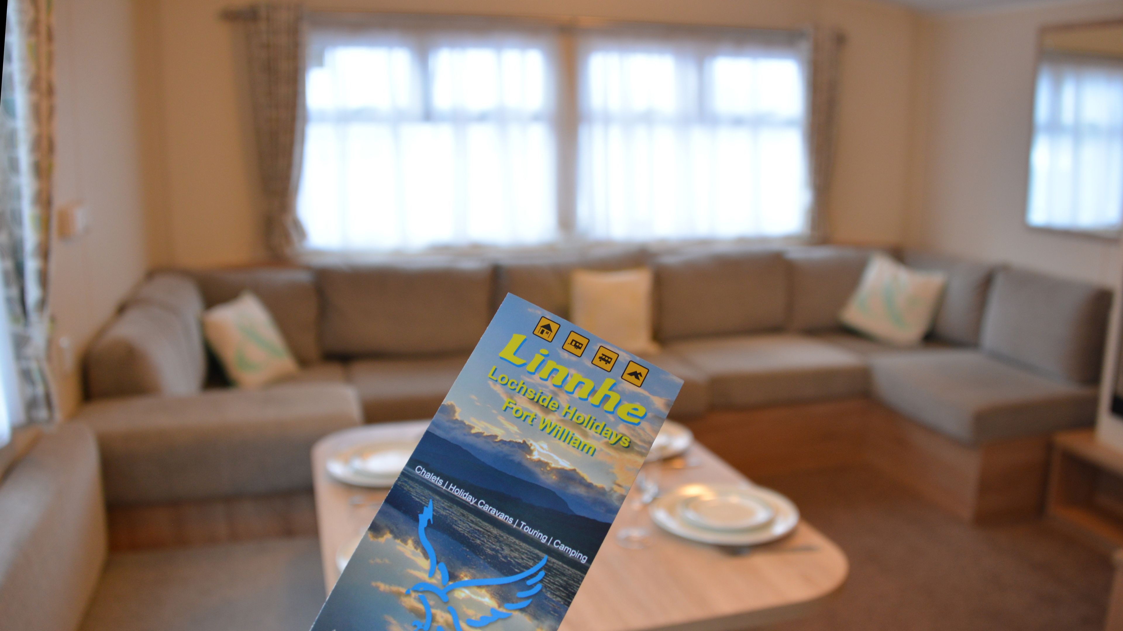 Un folleto con el título 'Linnhe Lochside Holidays Fort William' sobre una mesa en un salón con un sofá y una ventana al fondo.