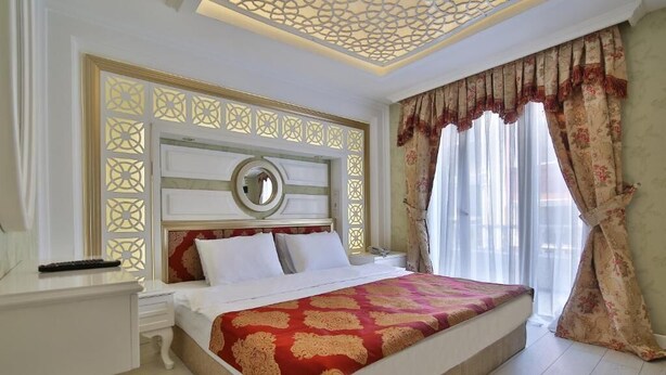 Une chambre d’hôtel avec un grand lit, une table de chevet, une commode, un plafond décoratif et une fenêtre avec des rideaux.