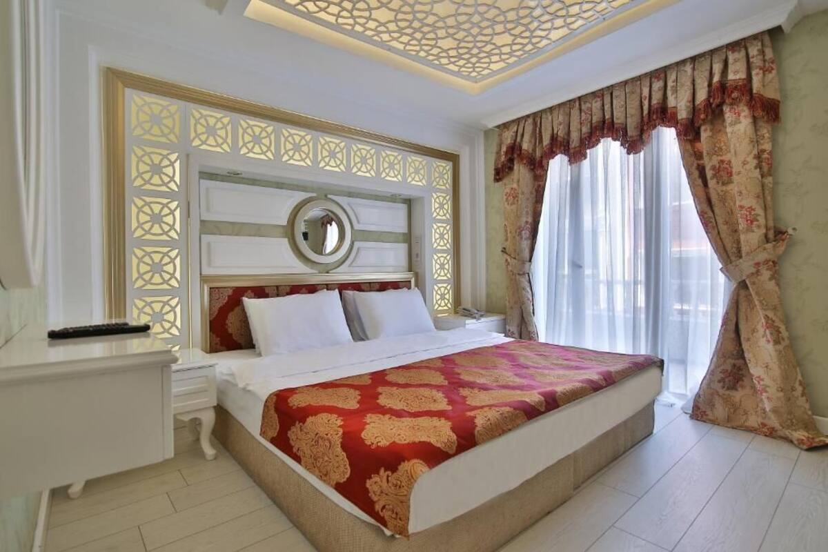 A hotel room with a large bed, a bedside table, a nightstand, a decorative ceiling, and a window with curtains.