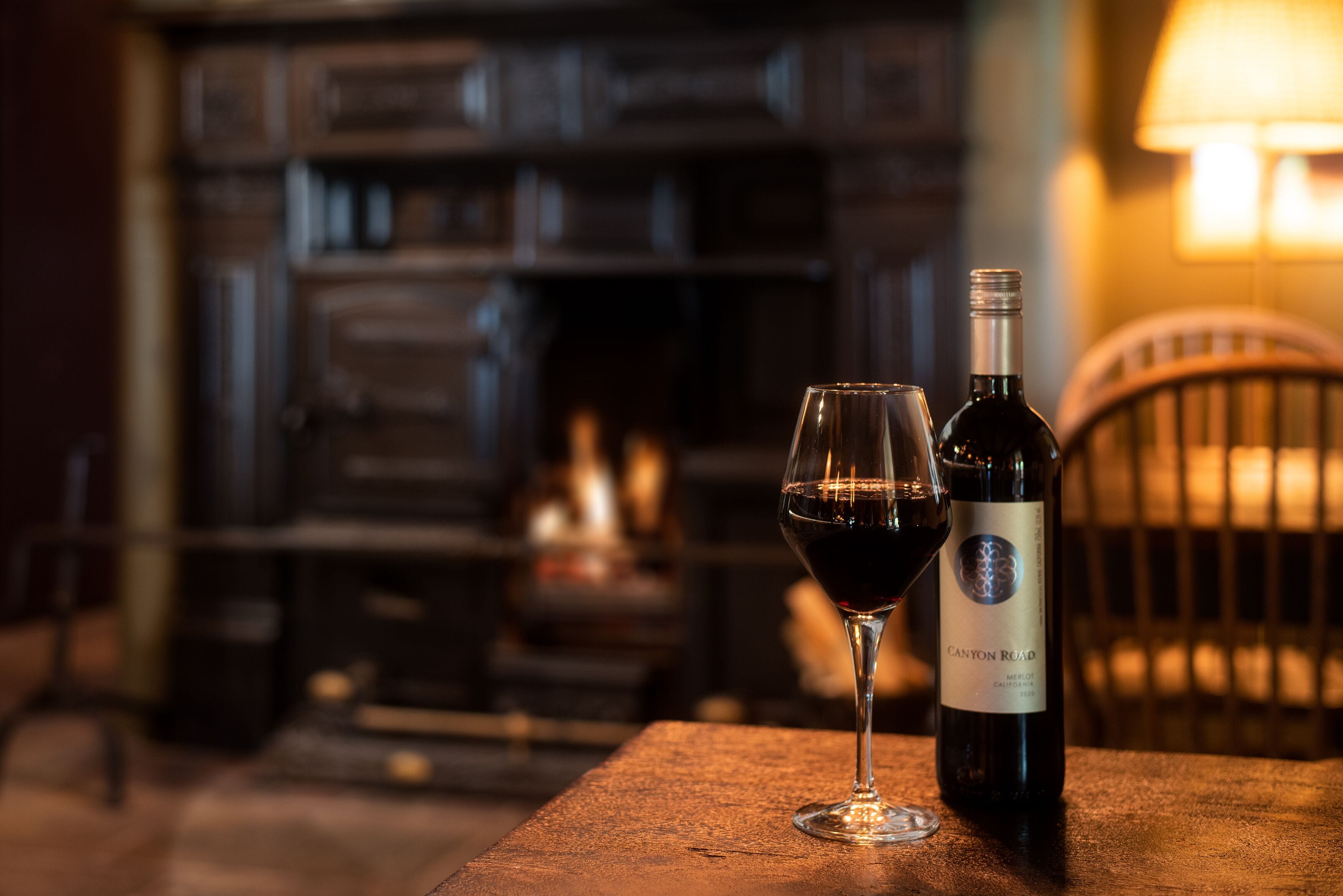 A bottle of wine and a glass on a wooden table with a blurred background of a room with wooden furniture and a lamp.