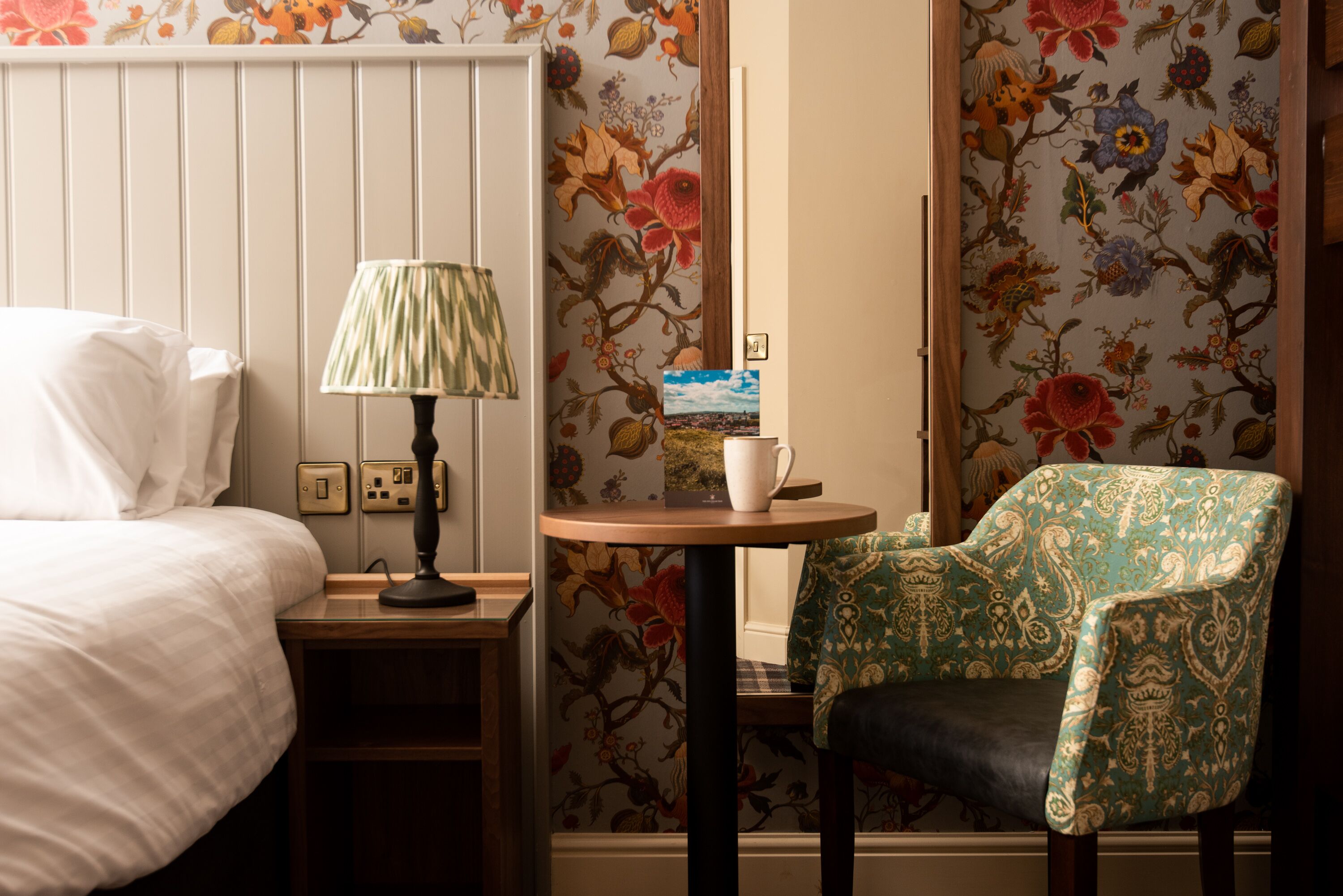 A hotel room with a bed, a nightstand with a lamp, a small table with a cup and a book, and a chair with floral wallpaper.