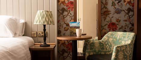 A hotel room with a bed, a nightstand with a lamp, a small table with a cup and a book, and a chair with floral wallpaper.