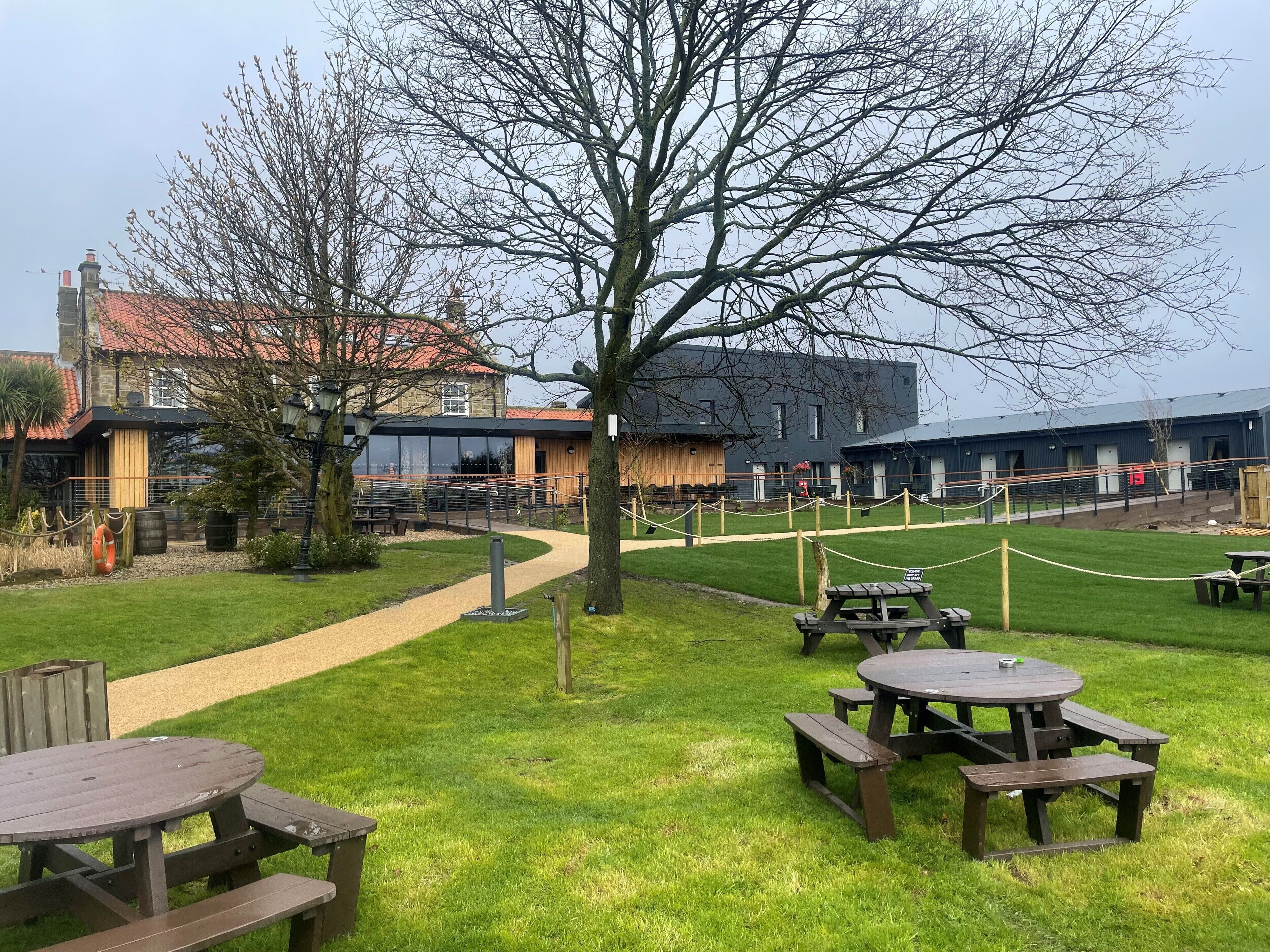 A grassy outdoor area with picnic tables and a pathway leading to a building with a red-tiled roof.
