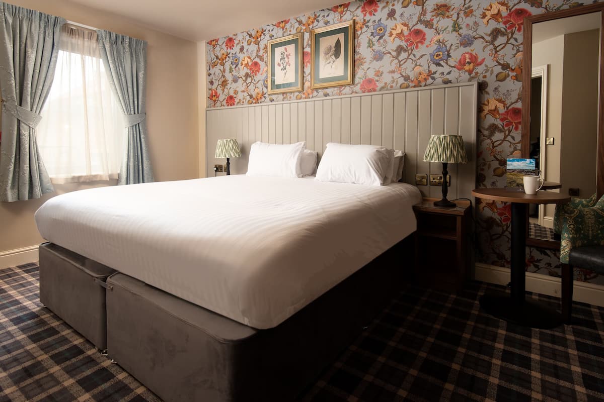 A hotel room with a large bed, a dresser, a nightstand with lamps, floral wallpaper, and a checkered floor.