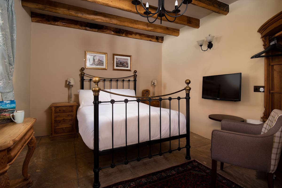 A bedroom with a four-poster bed, a television, a wooden nightstand, a chair, and a small table with a cup.