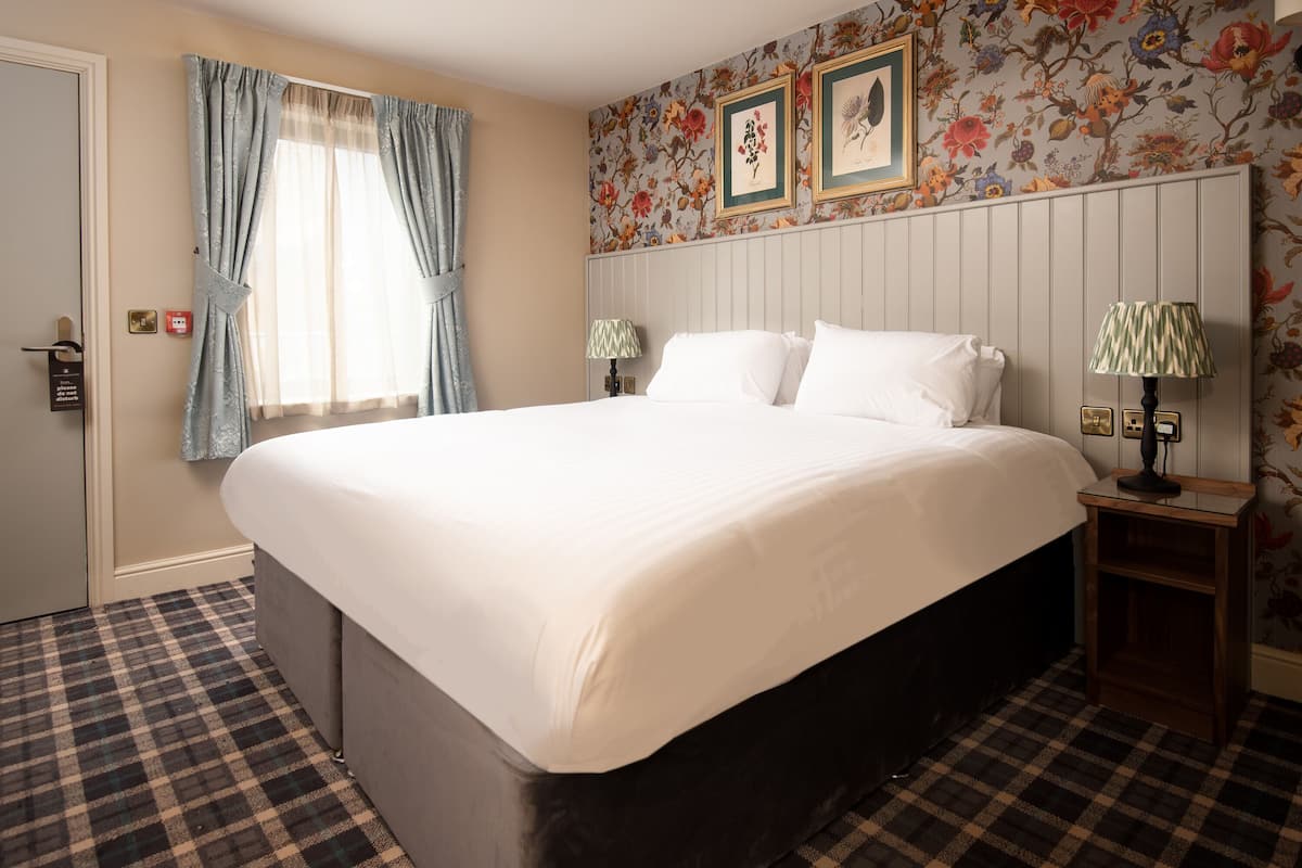 A large bed with a white duvet in a room with floral wallpaper, checkered floor, and two bedside lamps.