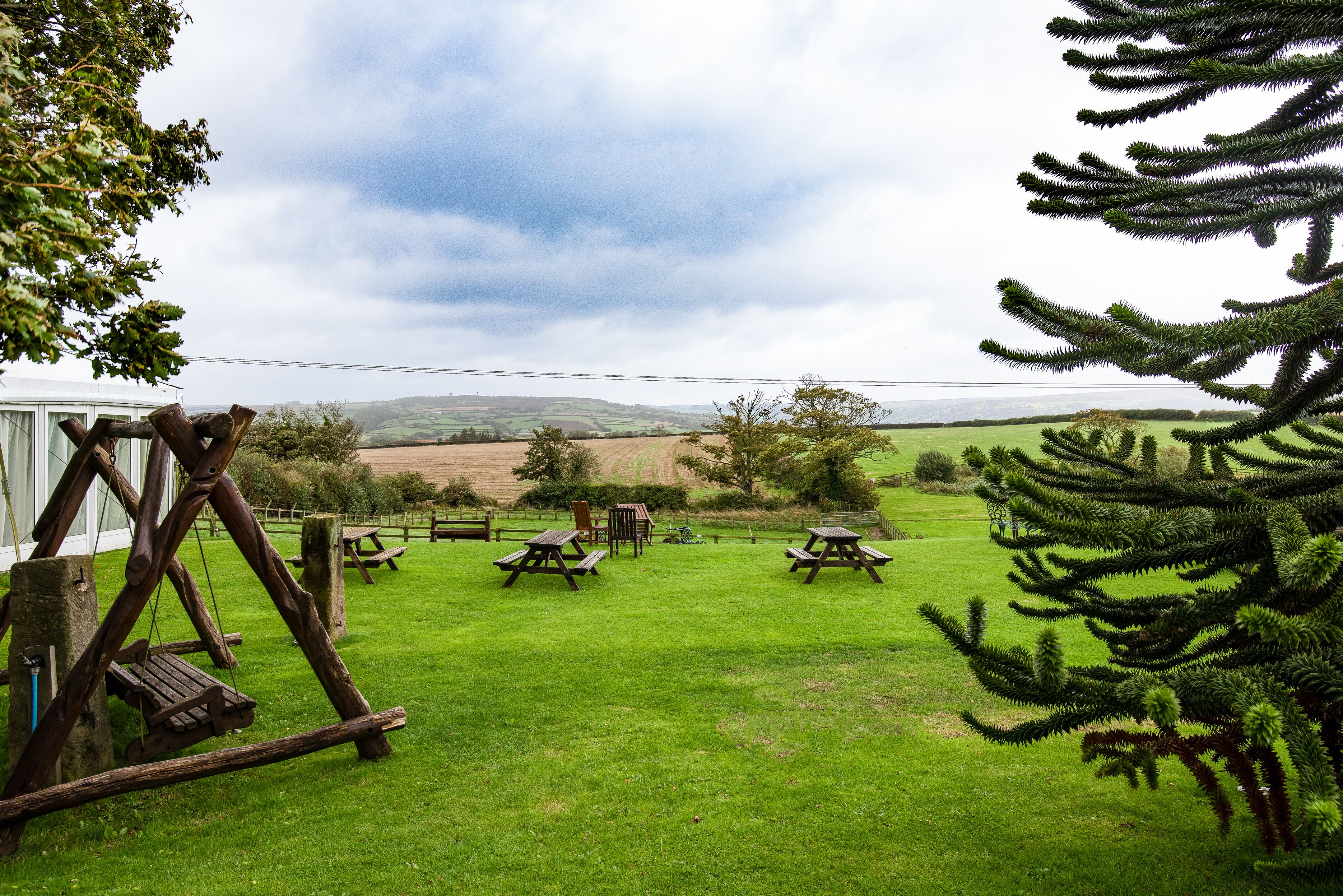A grassy outdoor area with picnic tables and a swing set, surrounded by trees and a view of distant hills.