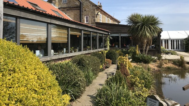 A building with a red-tiled roof, large windows, and a covered outdoor area with plants and a pond. - The Stables Whitby (Whitby)