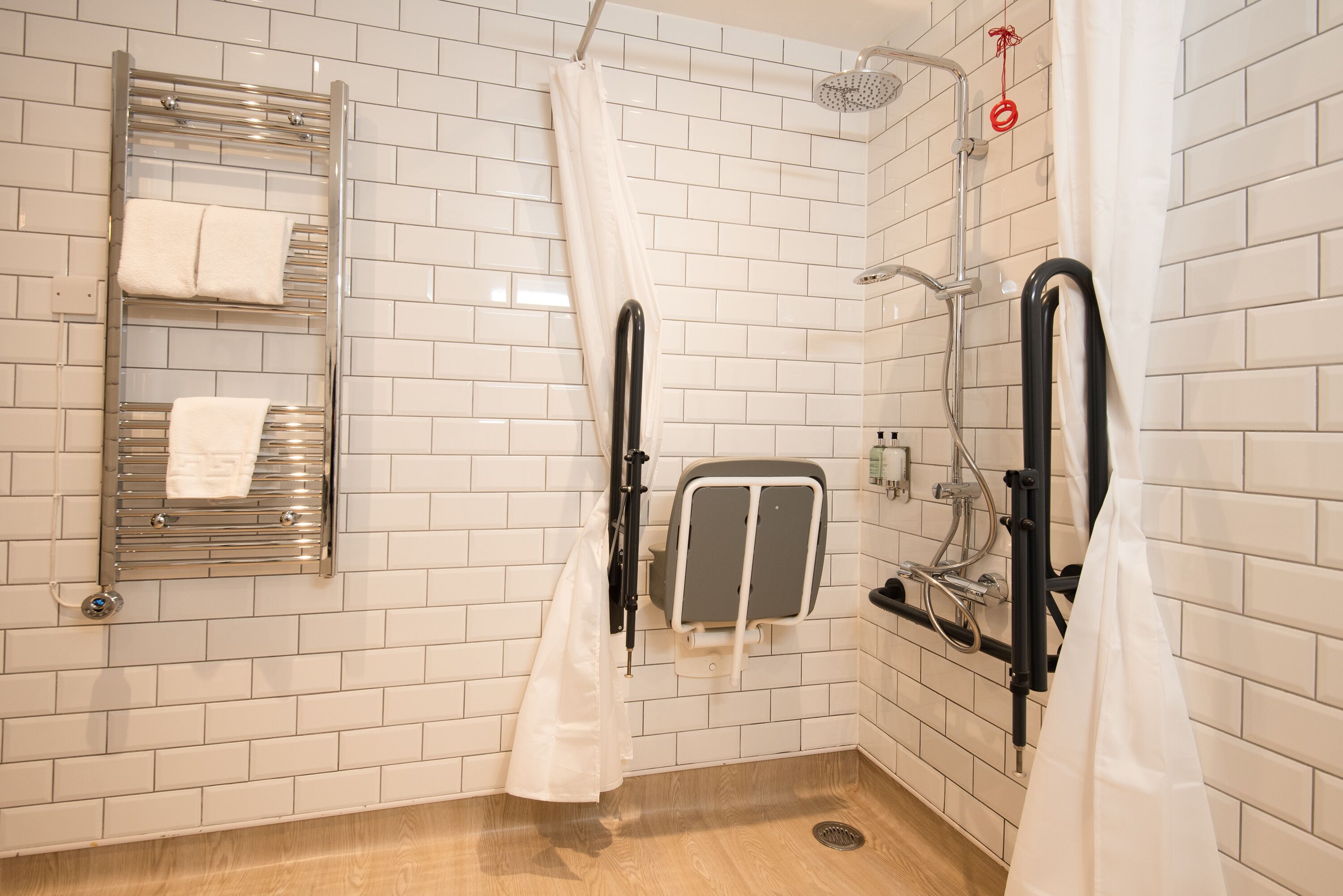 Un baño con paredes de azulejos blancos, ducha con cabezal de ducha manual, toallero y secador de manos.
