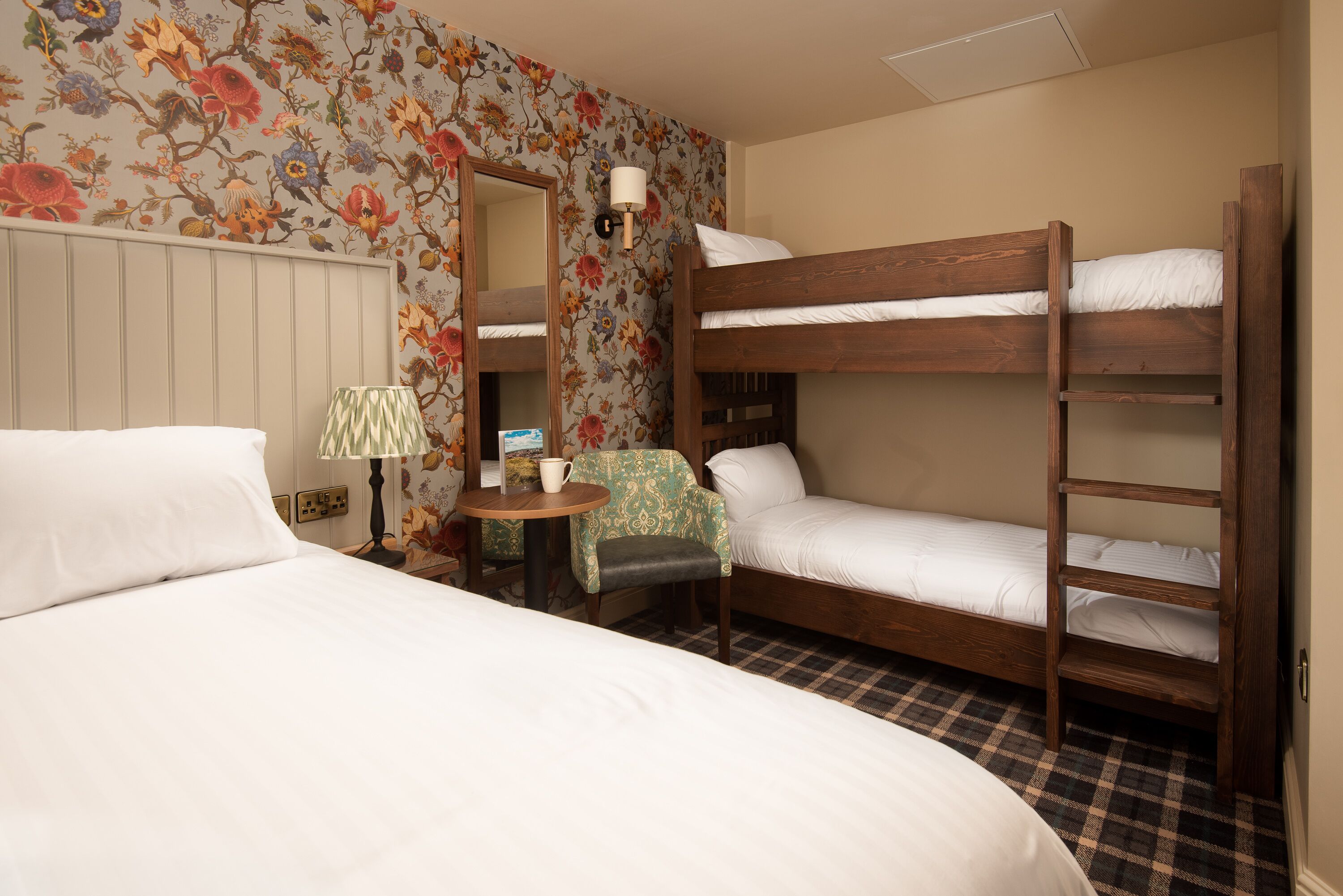 Habitación de hotel con una cama grande, una cama individual en literas, una silla, una mesa con lámpara y papel tapiz con motivos florales.