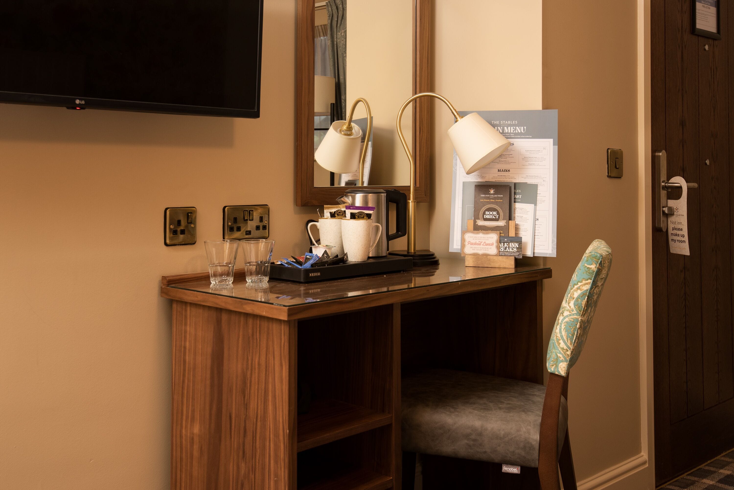 A hotel room with a wooden desk, a chair, a television, a mirror, and a menu board.
