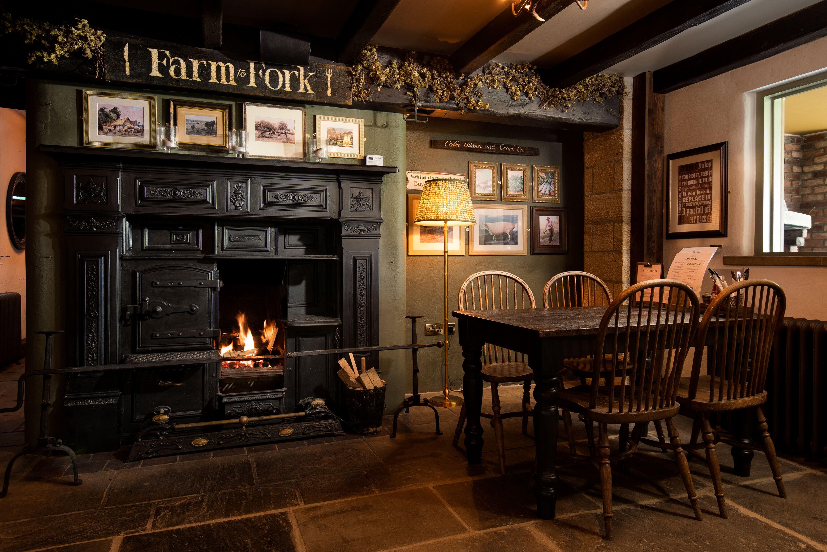 A fireplace with a sign above it reading "Farm to Fork." There are framed pictures on the wall and a table with chairs in front of the fireplace.